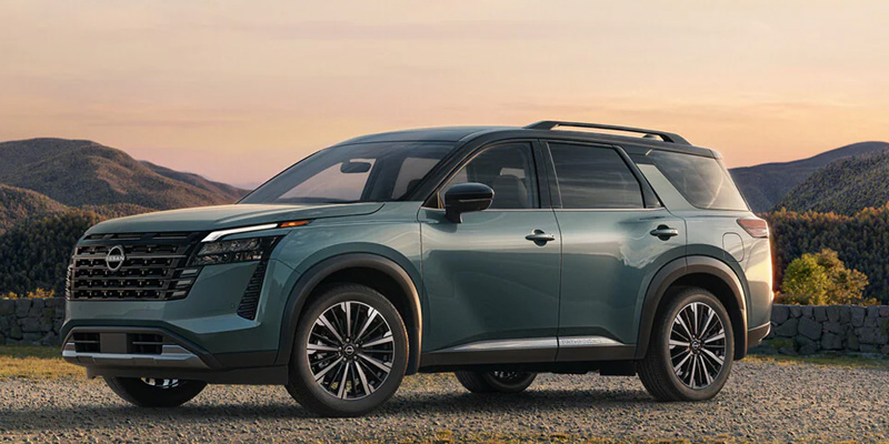 Green Nissan Pathfinder parked on gravel with hills at sunset.