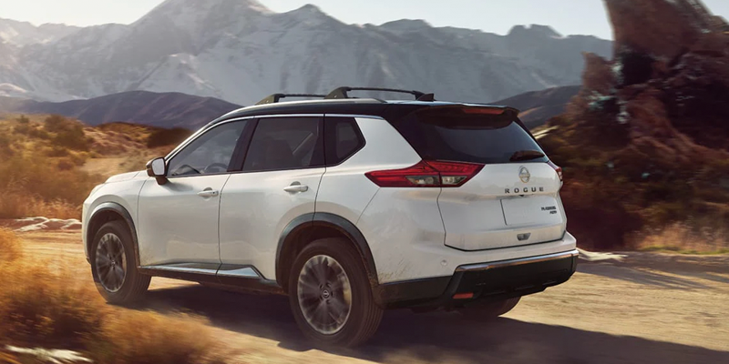 White Nissan Rogue driving on a dusty trail with mountains and desert terrain in the background.